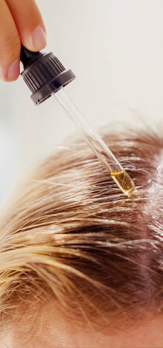 Immagine della riga centrale del cuoio capelluto, una mano tiene i capelli, l'altra sta applicando con la pipetta la fiala sul cuoio capelluto